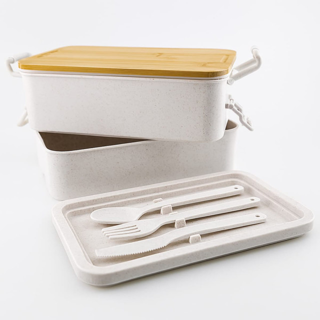 A dual-compartment lunch box with a yellow lid, featuring white utensils and an inner tray. It has a logo on the side.