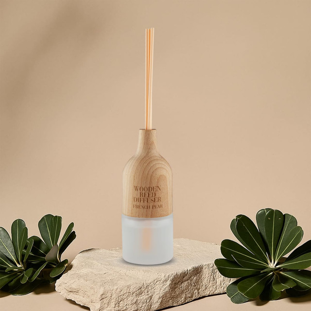 A wooden reed diffuser with a light wood top and a frosted glass base, displayed on a stone surface with greenery.
