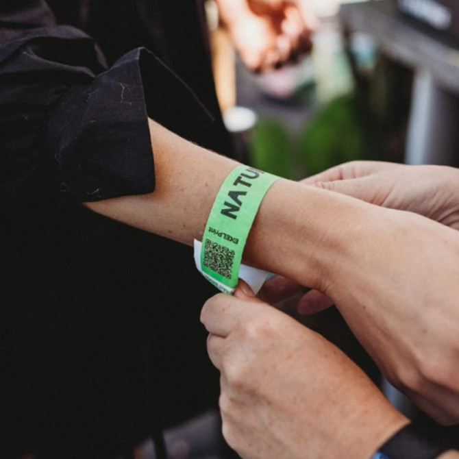 A green Tyvek adult wristband being fastened on an arm, featuring a logo and a QR code.