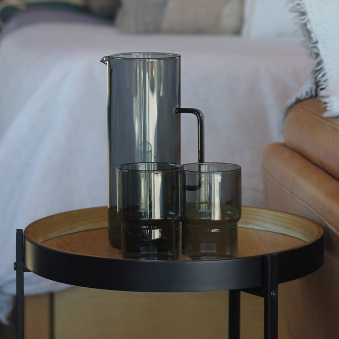 A drink bottle and two glasses in smoky grey, displayed on a wooden side table with a black frame.