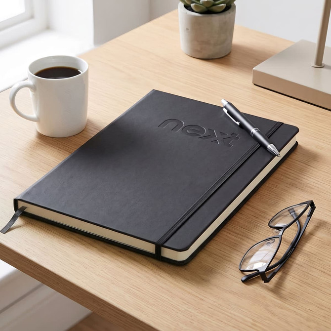 A black A4 daily diary on a wooden desk, accompanied by a coffee cup, a pen, and reading glasses.