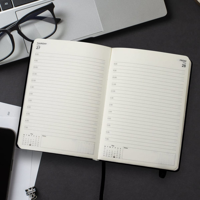 An open A5 daily diary with blank pages on a desk, alongside glasses and a laptop, featuring a black cover.