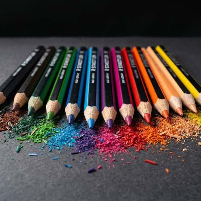 A set of 12 coloured pencils in various shades, arranged over colourful pencil shavings and debris. The set includes a logo.