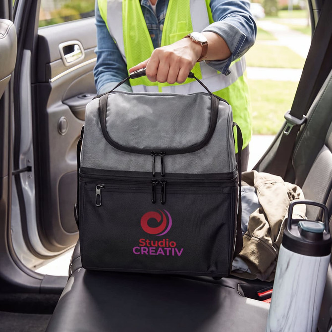 A grey cooler bag with a logo is on the front seat of a car, alongside a drink bottle and folded clothing.
