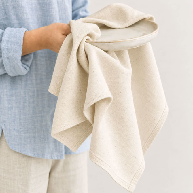 A person holds a large, cream cotton tea towel draped over a light ceramic plate. The fabric is textured and natural.