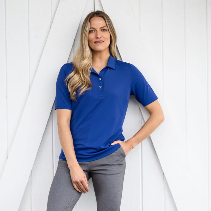 A woman in a blue polo shirt stands against a white wall, wearing grey pants and posing confidently.