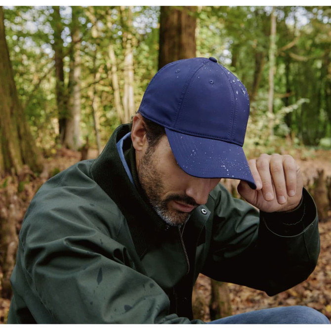 A navy cap worn by a man sitting in a forest, with water droplets on its surface. The background features trees and foliage.