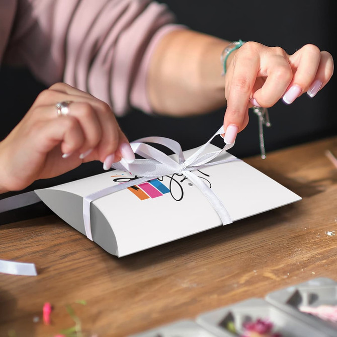 A medium white pillow box with a website logo, being tied with a white ribbon by a person.