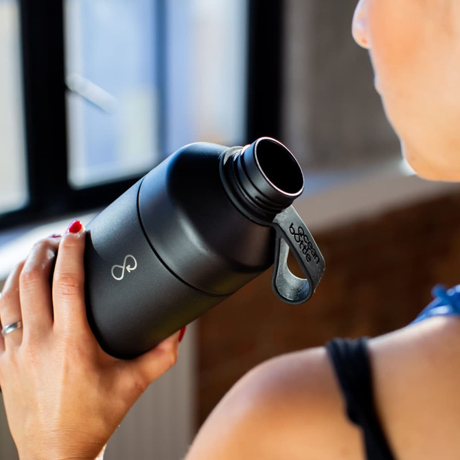 A drink bottle in matte black with a wide mouth and a handle, held by a person in a fitness setting.