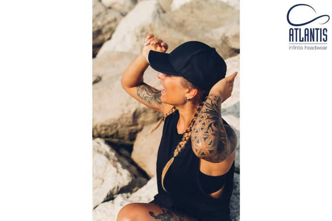 A black cap is worn by a woman with tattoos, seated on rocky terrain. The cap features a branding logo.