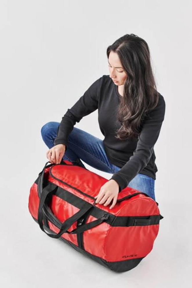 A medium red waterproof gear bag with black straps and a logo, held by a person seated on the floor.