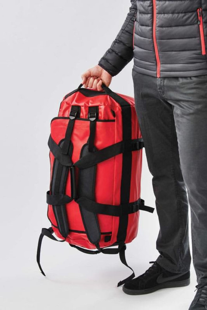 A red waterproof gear bag with shoulder straps, held by a person wearing a black puffer jacket and grey pants.