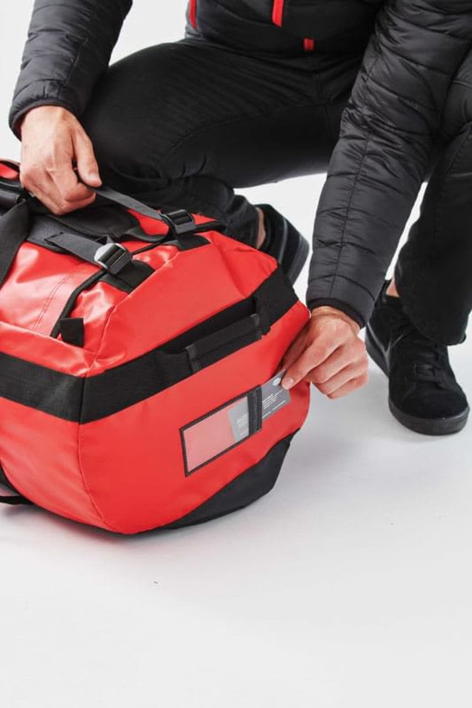 A medium waterproof gear bag in red and black with adjustable straps and a tag pocket. Includes a logo.