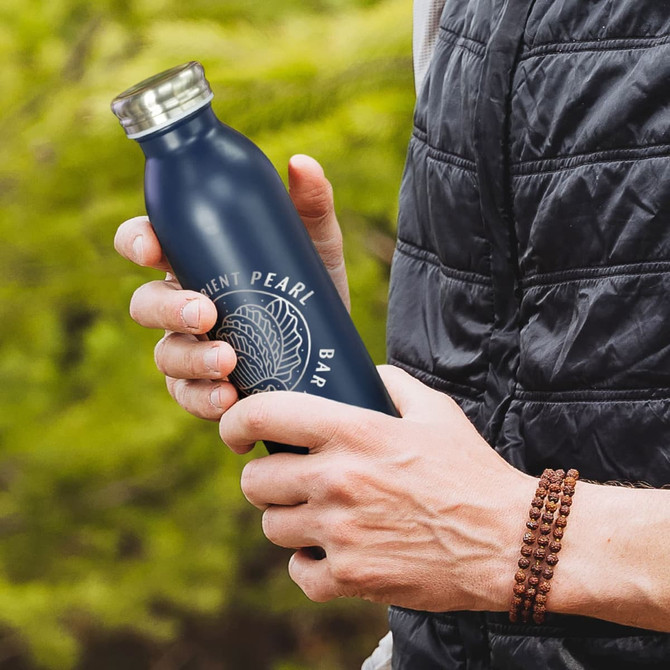 A drink bottle in navy blue held by a person, featuring a logo on the front against a natural background.