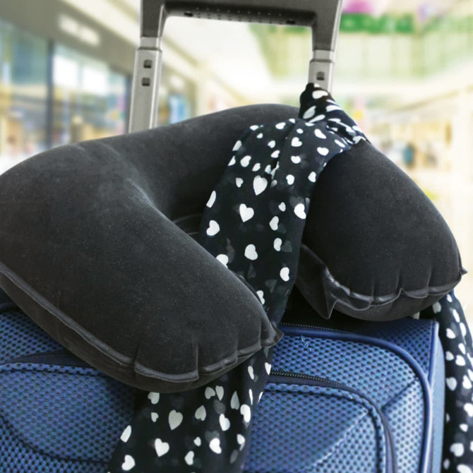 A black neck pillow resting on a suitcase, with a patterned scarf draped over it. The background is blurred with vibrant colours.