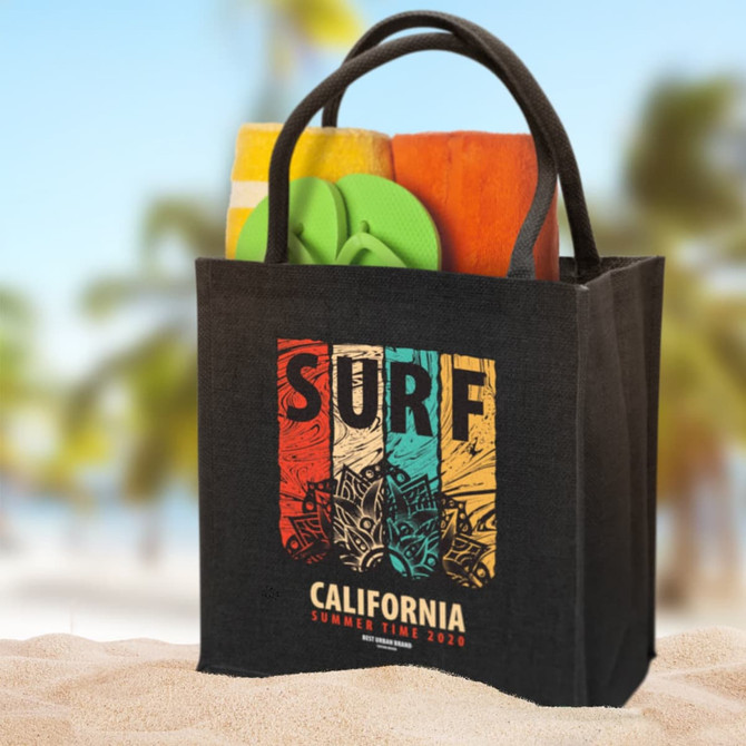A black jute tote bag holds a towel, flip-flops, and other beach items. The bag features a logo on the front.