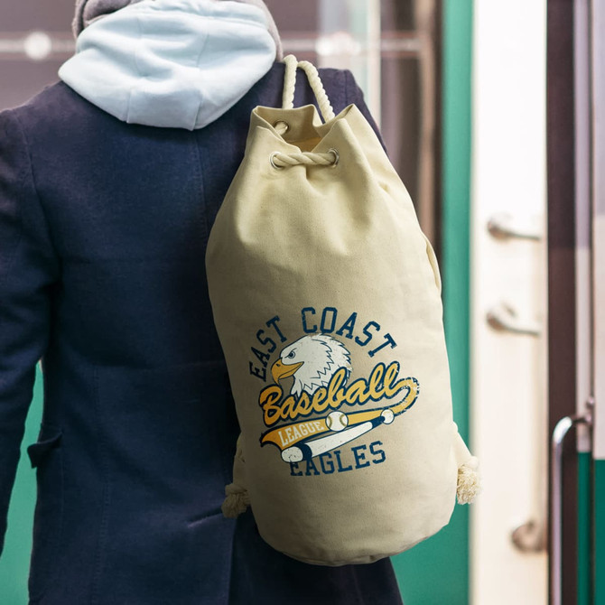 A beige canvas barrel bag with a drawstring and an eagle logo. It is slung over a person's shoulder.
