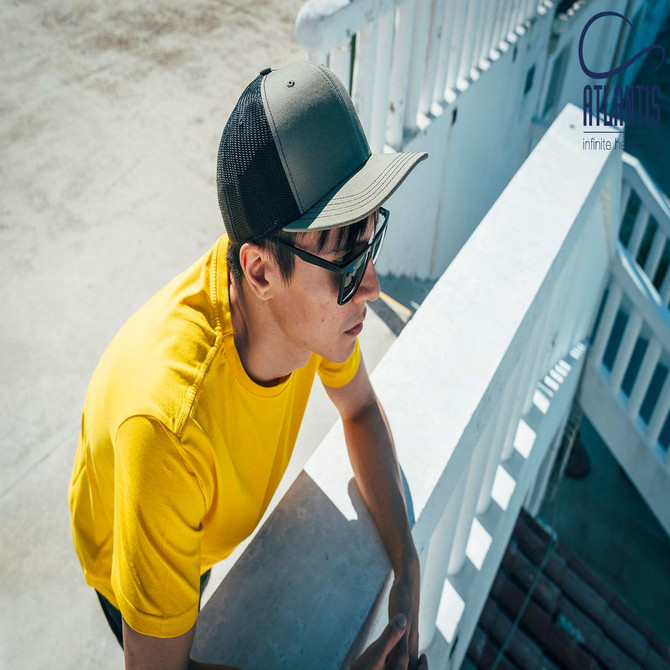 A man wearing a grey trucker cap and a yellow shirt, leaning against a white railing. The cap has a logo.
