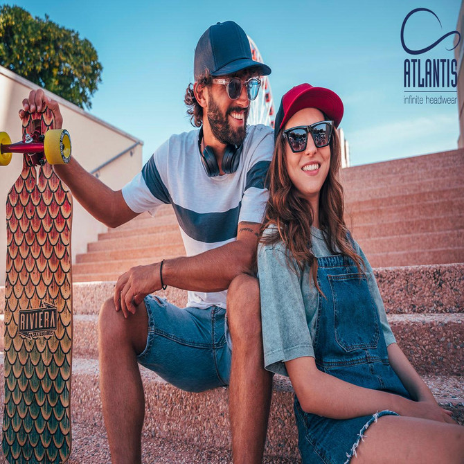 A trucker cap featuring a dark grey front and red mesh back, alongside a skateboard and two people sitting on steps.