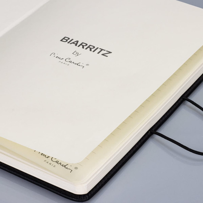 A black notebook with a ribbon bookmark, featuring plain cream pages and branding on the inside cover.