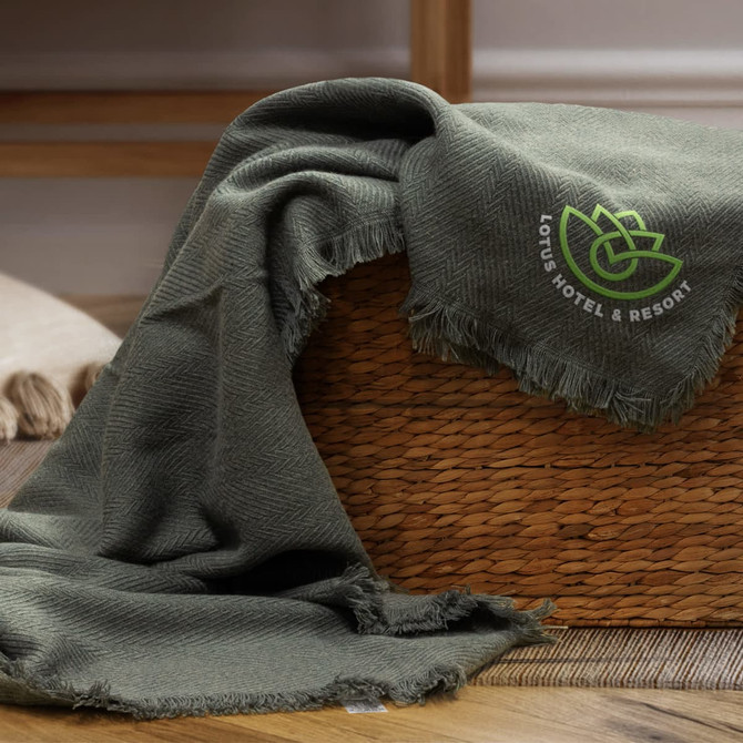A green herringbone throw blanket draped over a woven basket, featuring a small logo.