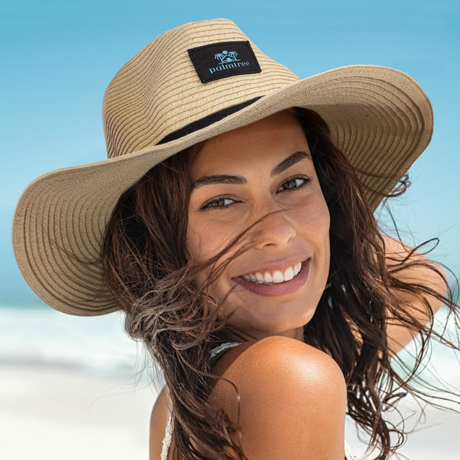 A wide brim hat in tan, featuring a logo, worn by a smiling woman with wavy hair against a beach backdrop.