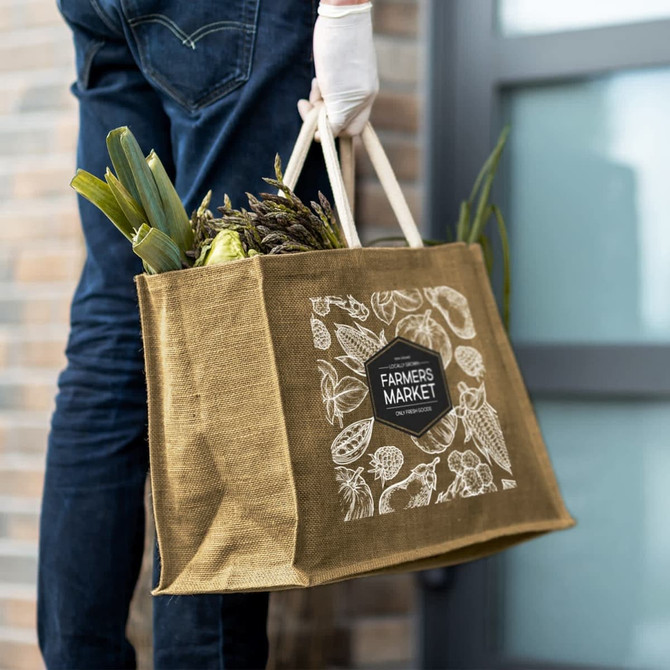 A jute tote bag in brown, featuring a logo and filled with fresh vegetables, held by a person wearing gloves.