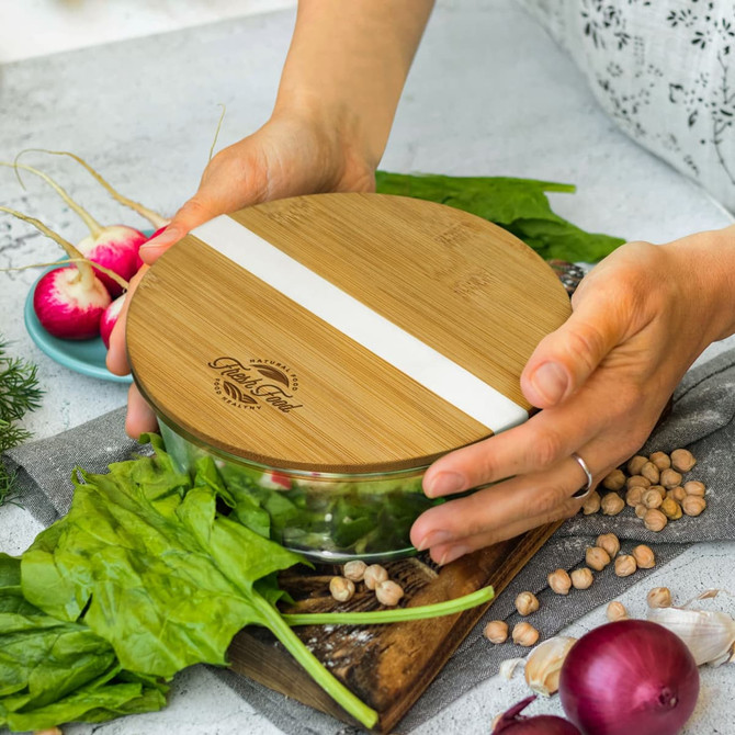 A glass food container with a bamboo lid, featuring fresh vegetables and herbs on a wooden cutting board.