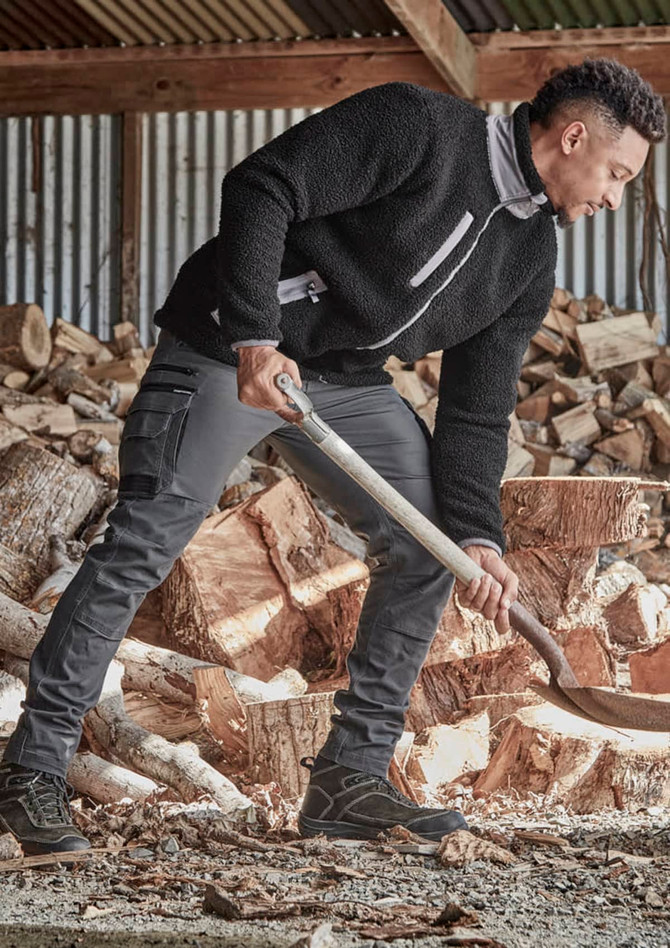 A man wears a black full zip sherpa fleece jumper with grey accents, standing in front of stacked logs.