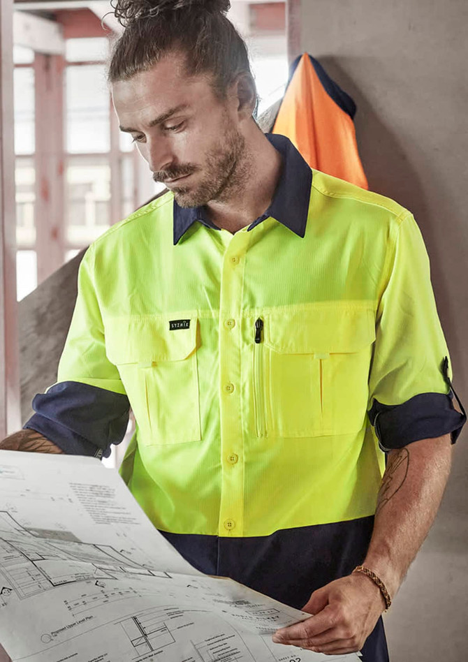 A man in a high-visibility yellow and navy long sleeve work shirt examines a blueprint in an indoor setting.