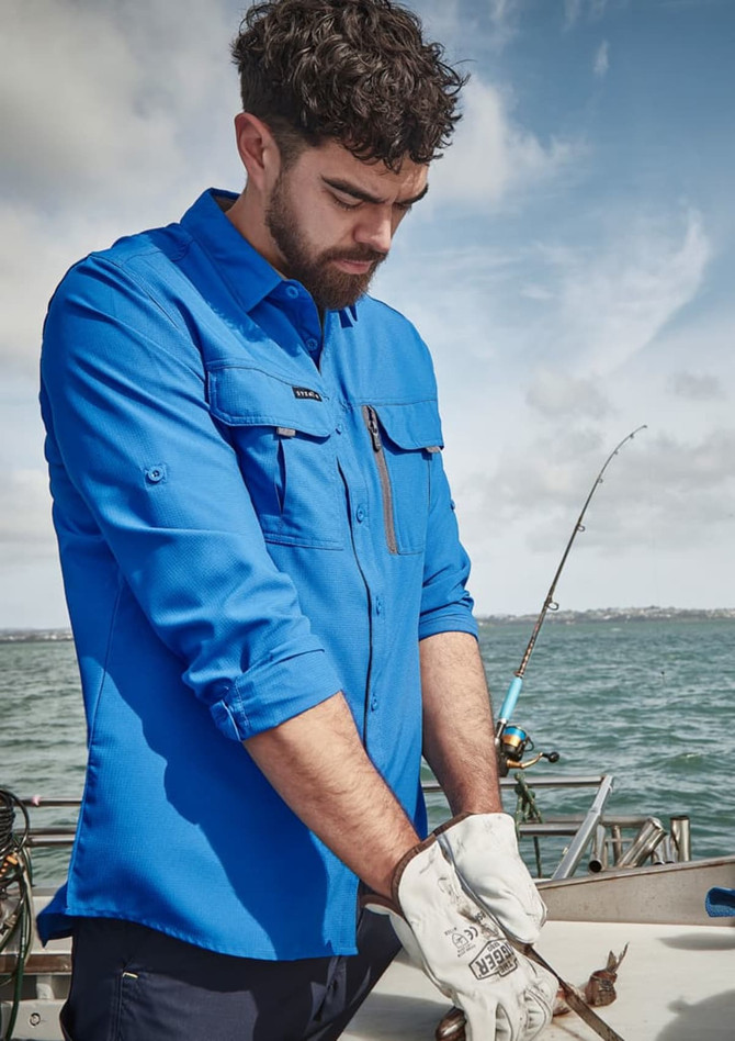 A man wearing a long sleeve blue button-up shirt, equipped with gloves, standing on a boat by the water.