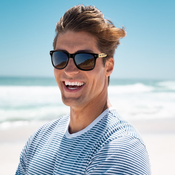 Bamboo sunglasses with a dark frame, worn by a man with a striped shirt, smiling against a beach backdrop.