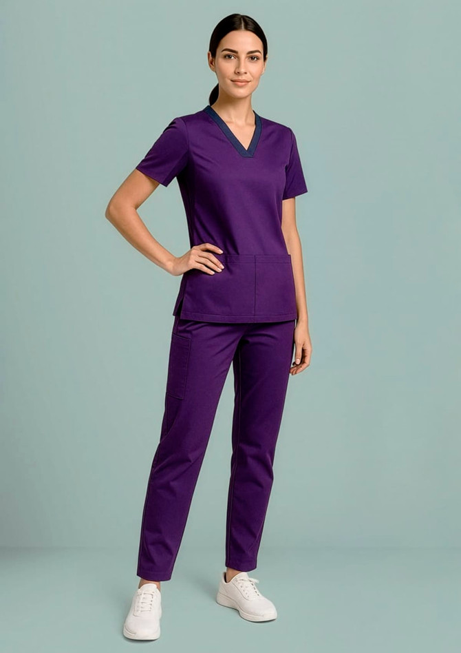 A woman wearing a purple women's Riley Stretch scrub top paired with matching pants and white sneakers, standing against a light background.