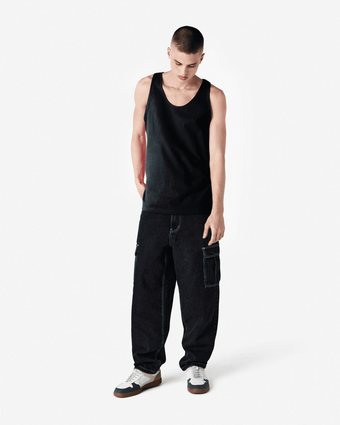 A male model wearing a black unisex fine jersey tank top paired with dark cargo pants and white sneakers.