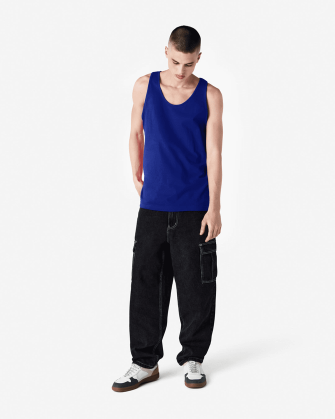 A blue unisex fine jersey tank top worn by a model with black cargo pants and shoes. The tank features a logo.