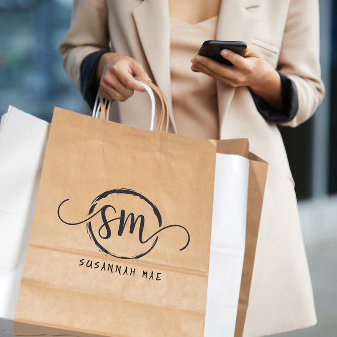 A medium brown paper bag with handles, featuring a logo and held by a person in a light-coloured jacket.