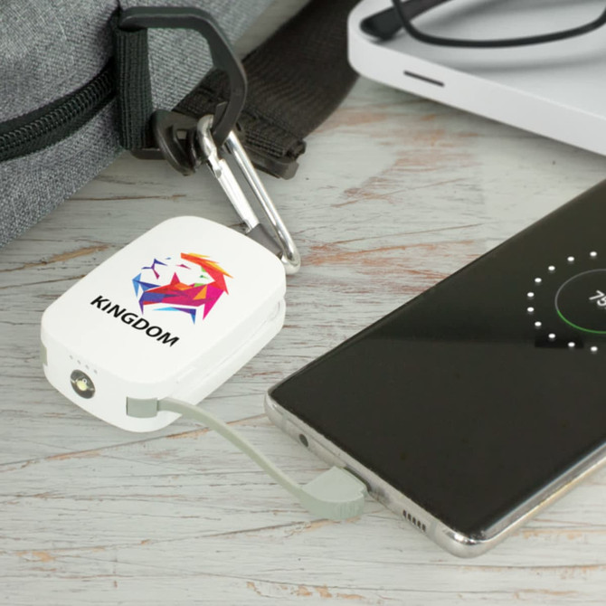 A white power bank with a logo, connected to a smartphone, placed on a grey surface beside a bag.