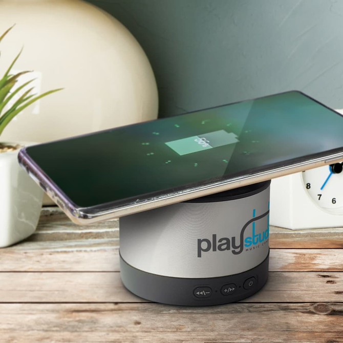 A wireless speaker charger in grey with a smartphone placed on top, featuring touch controls and a logo.