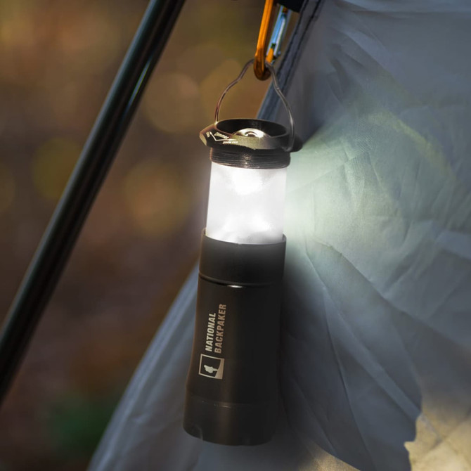 A black flashlight lantern hangs from a tent, emitting a bright white light. It features a loop for attachment and has branding.