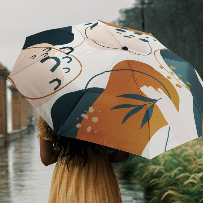 A compact umbrella with a full-colour design featuring warm tones and abstract shapes, held by a person in a yellow dress.