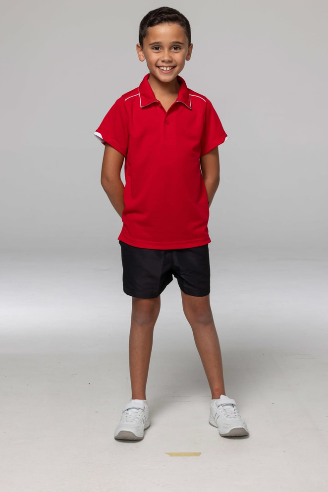 A boy wearing a red polo shirt and black shorts, standing with hands behind his back. The shirt features a logo.