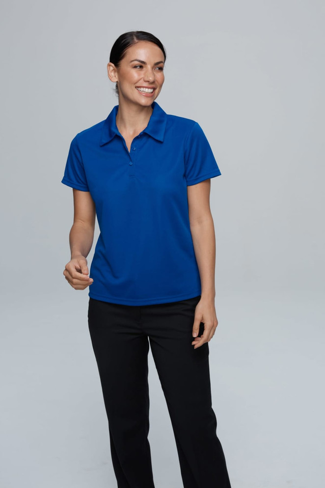 A woman in a blue polo shirt stands smiling against a neutral background, wearing black trousers. The shirt features a logo.