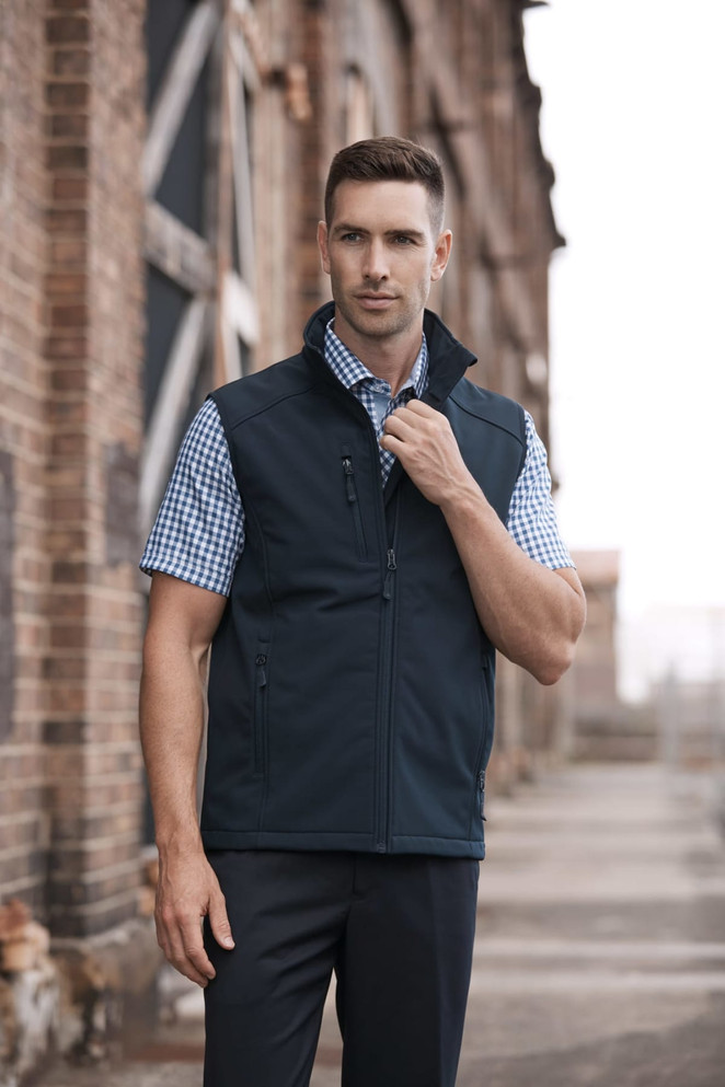 A man wearing a black vest over a checked shirt, standing outside near brick structures. The vest has a logo.