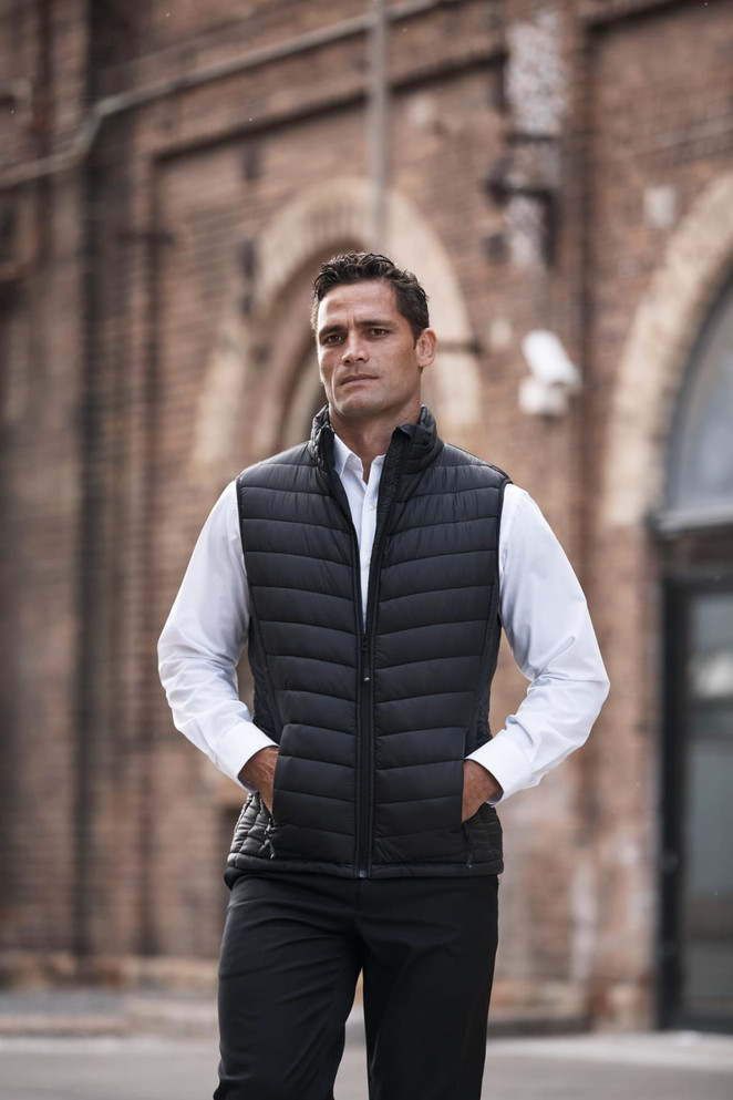 A black padded men's vest is worn over a white shirt, with the individual posing outdoors against a brick wall.