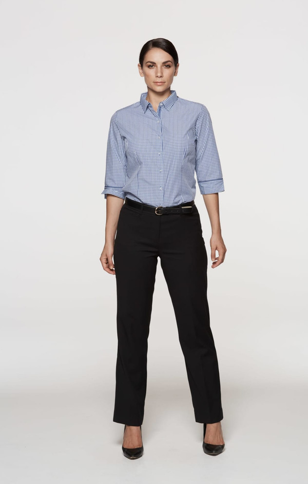 A woman stands in black pants and a blue checkered shirt, featuring rolled sleeves and a belt. The image includes branding.