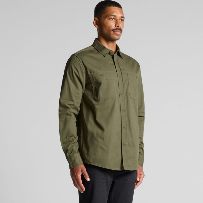 A man in an olive green work shirt, featuring two chest pockets and a button-up design, stands against a light grey background.