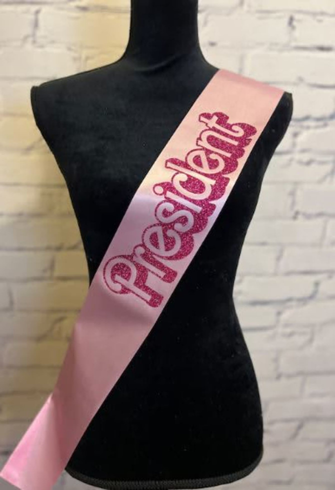 A pink satin sash with the word "President" in glittery lettering. It is displayed on a mannequin against a brick background.