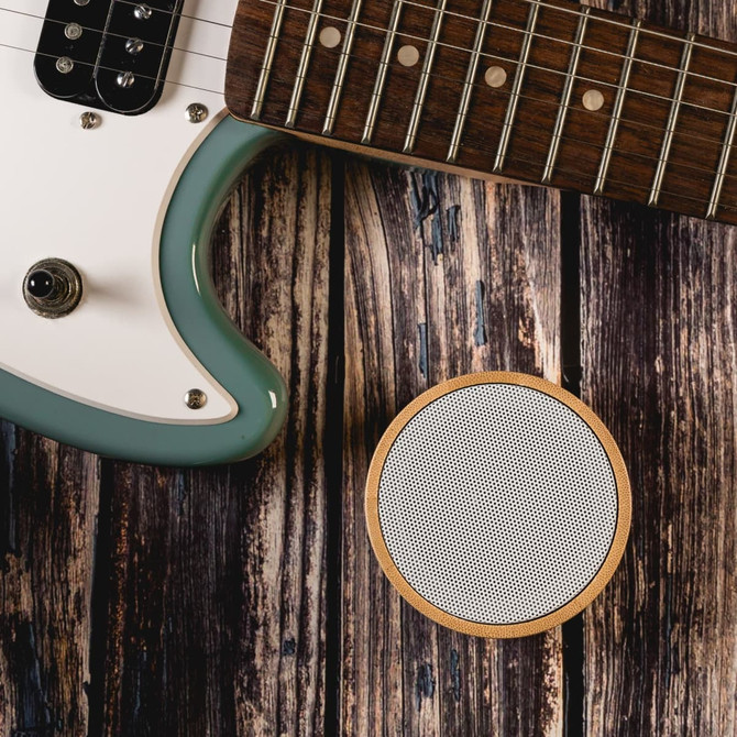 A round bamboo Bluetooth speaker in white, placed on a rustic wooden surface next to a green electric guitar.