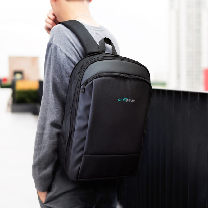 A black soft touch backpack with a logo, worn by a person, set against a cityscape backdrop.