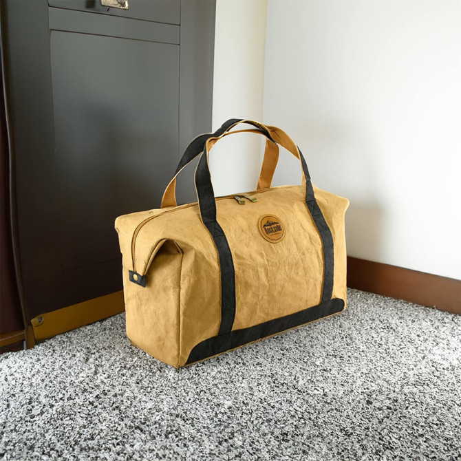A tan Kraft paper travel bag with black straps, featuring a logo, placed on a textured floor.